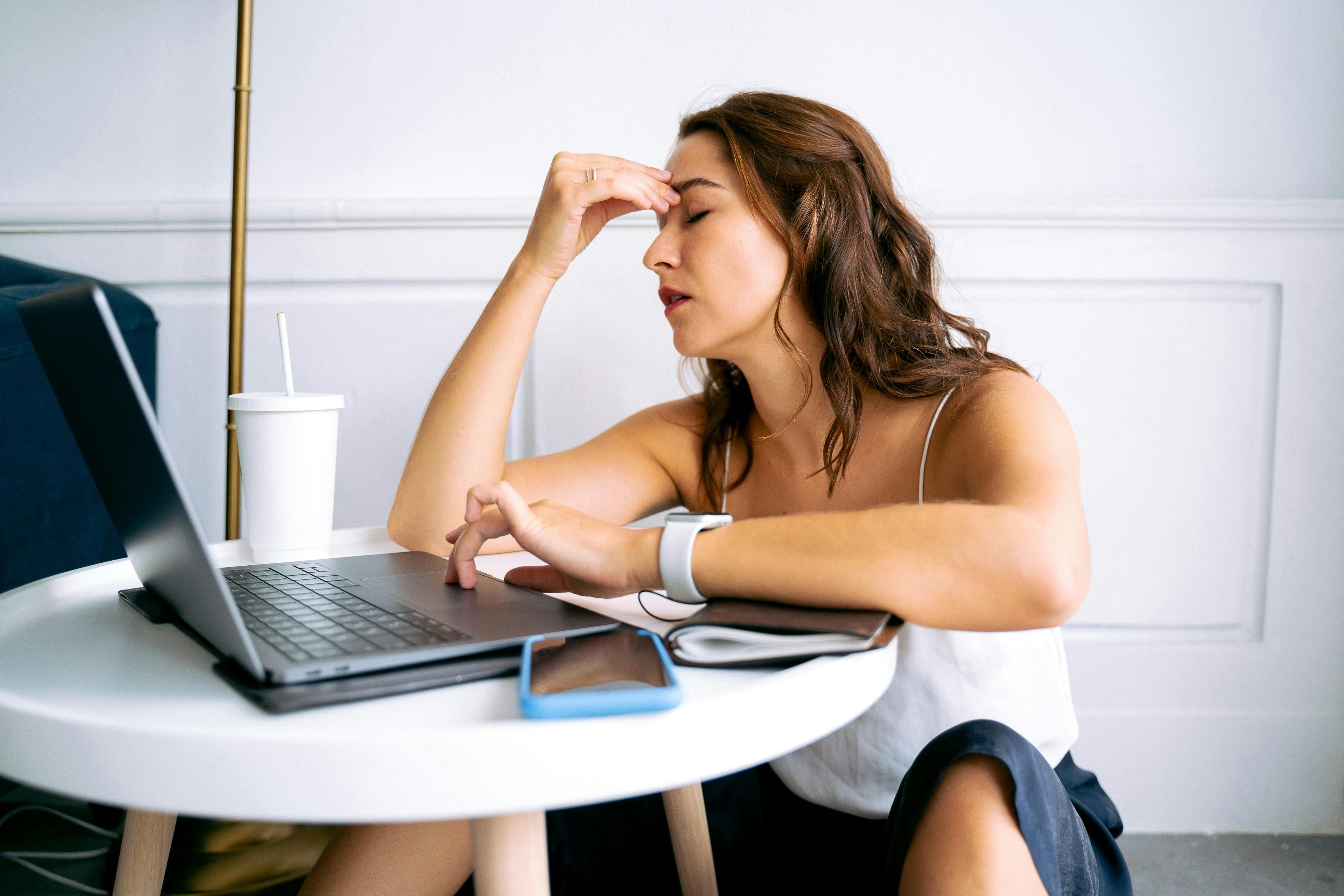 A woman sitting in front of her computer, holding her head due to a painful hormonal migraine triggered by poor sleep.